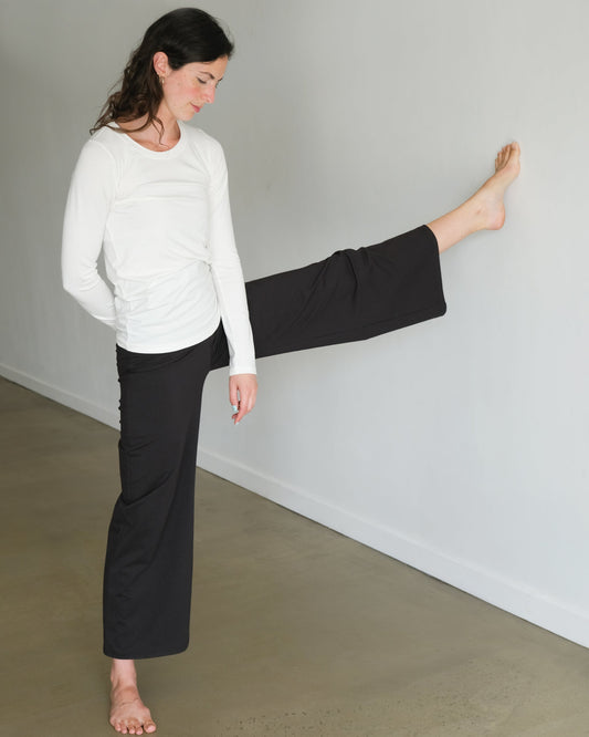 Woman in white sweater and black pants stretching against a plain wall.