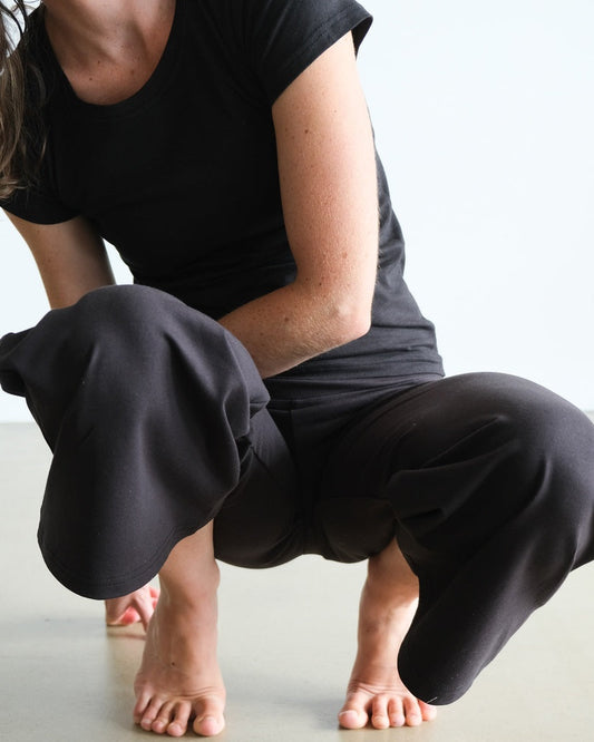 A person is crouching and demonstrating the flexibility of black wide leg trousers by pulling one leg behind them. The trousers appear to be made of a cotton-elastane blend and have a smooth, solid color with a foldable waistband.